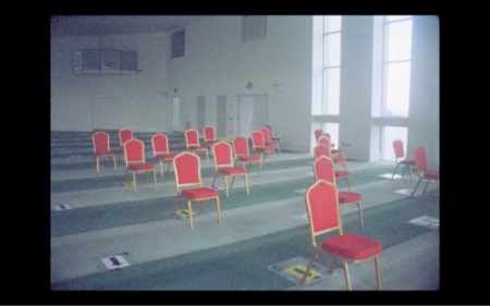 There are four rows of red chairs on a stripey carpet that alternates between light and dark green. There are a few signs laying on the floor with arrows pointing toward the same direction the chairs are facing. There are two large floor to ceiling windows letting in a lot of light, with a few other doors and windows further back.