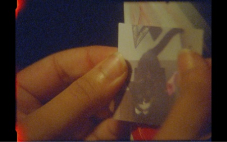 The film is about to burn out. Two hands are placing a small square sticker of a black and white kitten onto a red and white striped surface. The background is black.