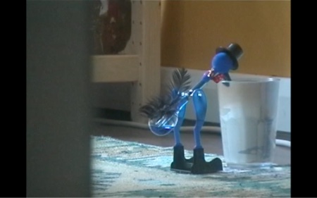 A blue drinking bird toy is hinged at the hips with its face in a glass of water, away from the camera. It is on the floor that has a green and white Turkish rug on it. The corner leg of a bookshelf can be seen in the background. The wall paper is yellow. There is a wire cable running along the bottom of it. Just under a quarter of the screen is obscured by a door.