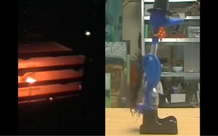 The screen is split down the middle. On the left side of the screen, the camera is looking through a gate with horizontal wires that has droplets of water hanging off of it. In the background, there is a glowing orange streetlight outside of a house. On the right side of the screen, the blue drinking bird toy is standing on a light brown wooden table facing the right. Its profile is fully visible. In the background there are a range of green toned paintings.